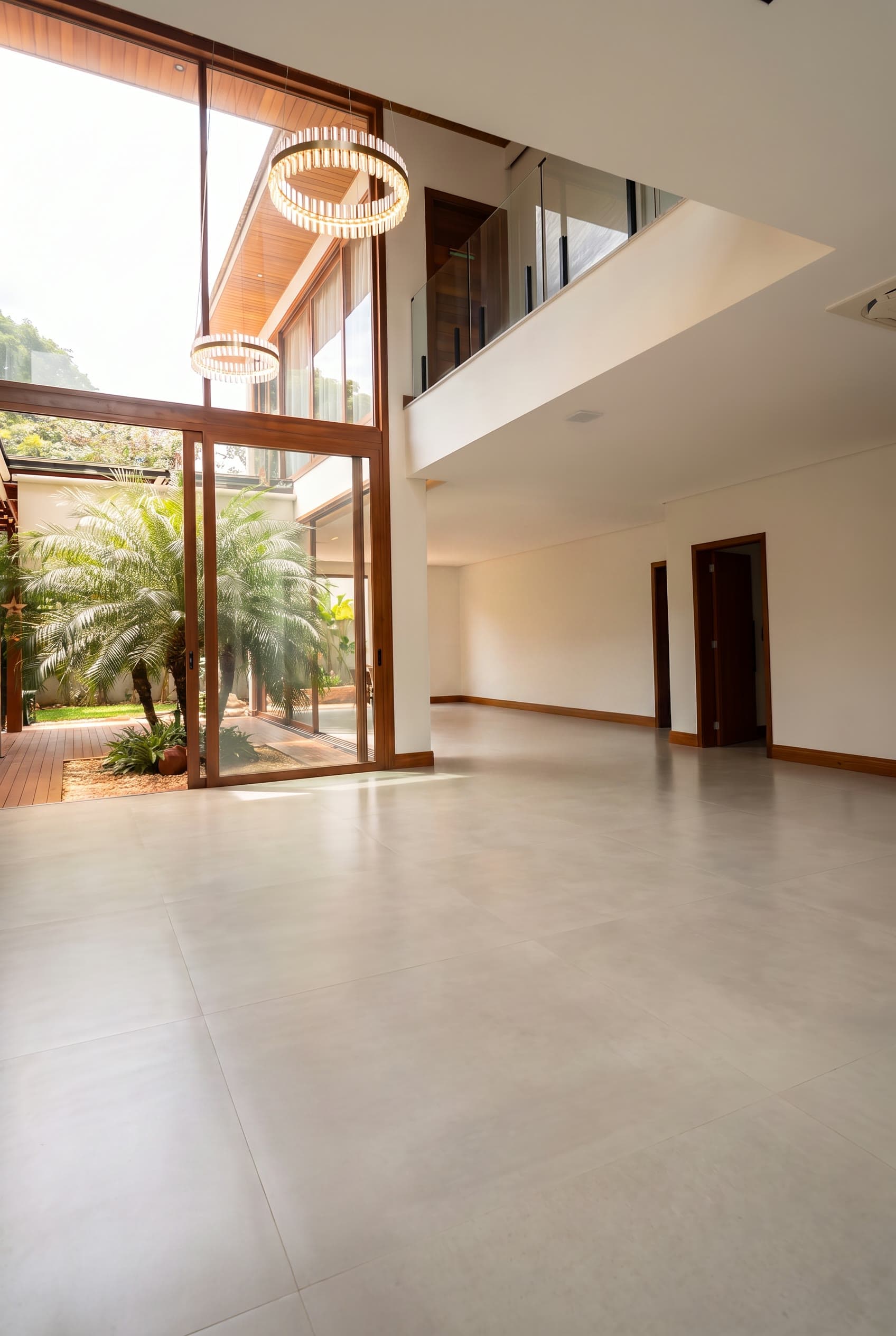 Sala vazia de pé-direito duplo, com esquadria de madeira, lustre circular e jardim com palmeira ao fundo