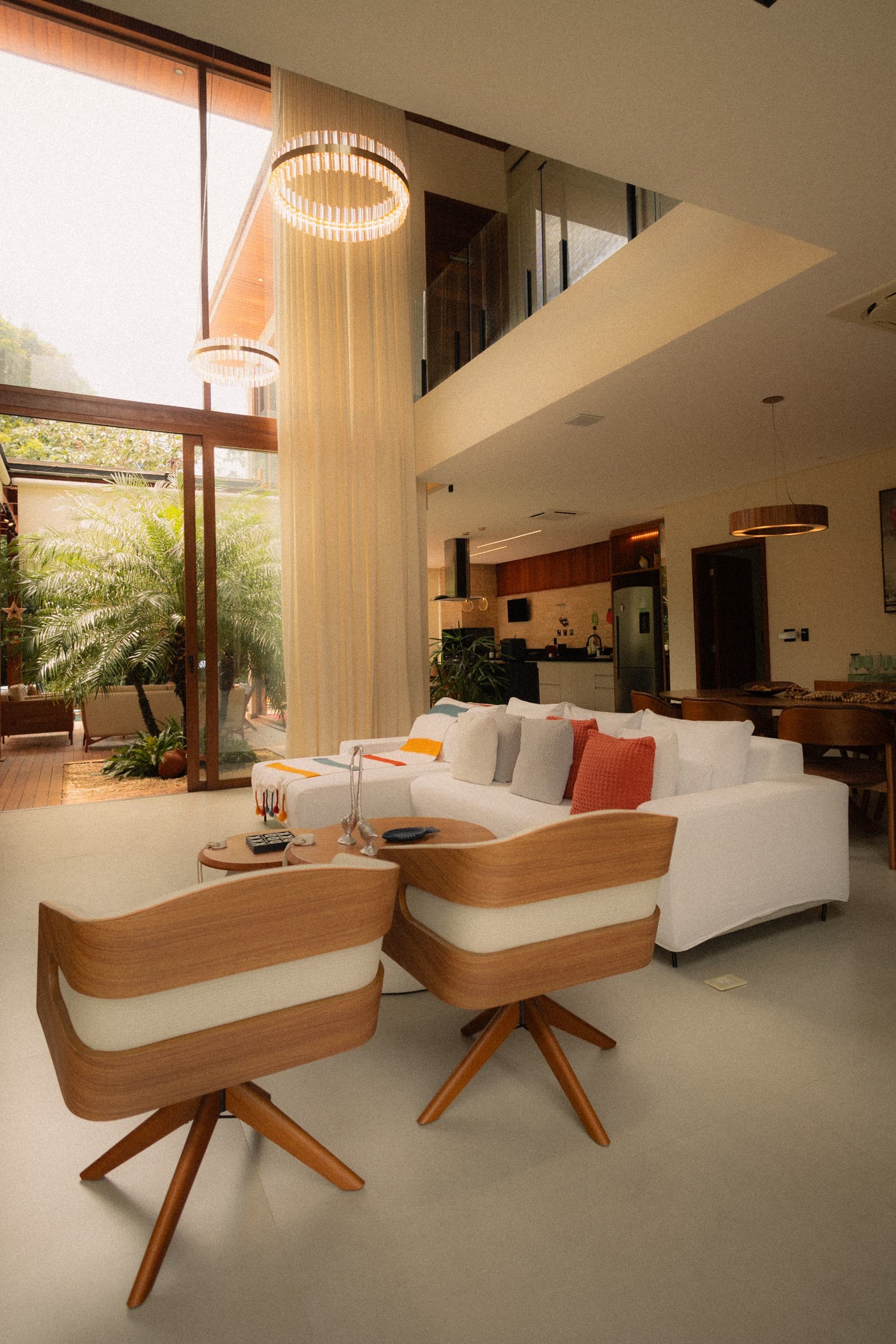 Sala com pé-direito duplo ambientada virtualmente, com sofá branco, duas poltronas giratórias de madeira e jardim ao fundo